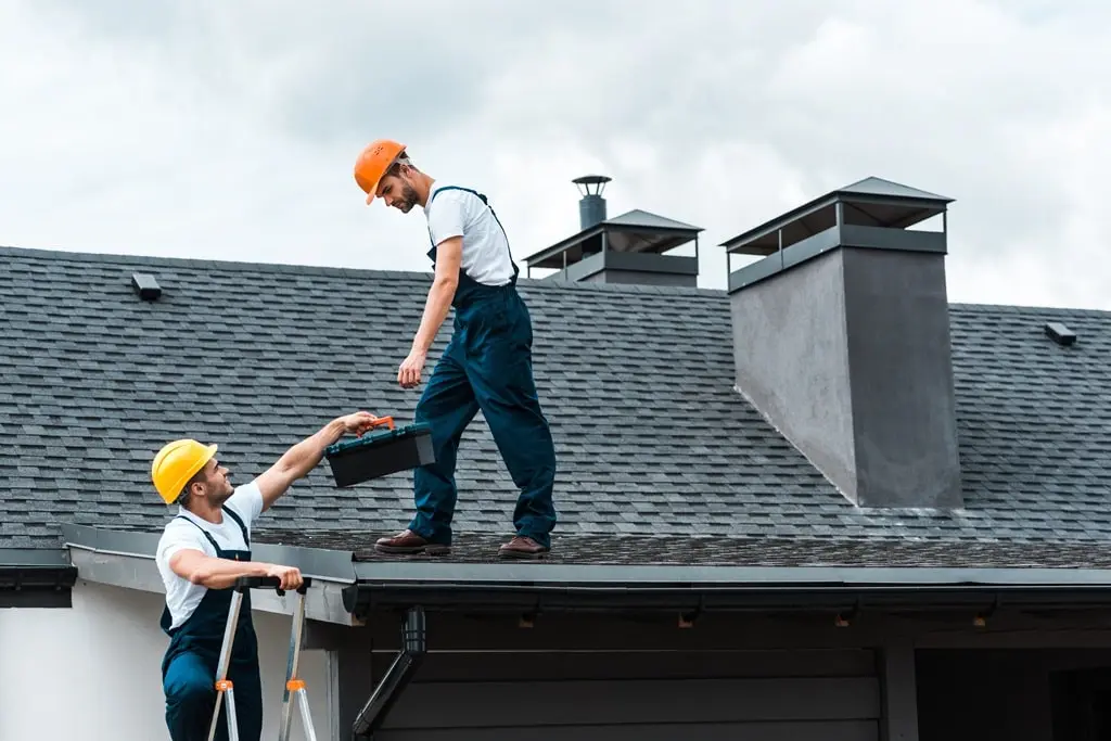 A handyman in a helmet standing on a ladder gives a toolbox to a colleague standing on a roof.