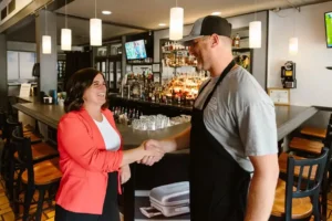 A businesswoman and a bar owner shake hands in front of the bar.
