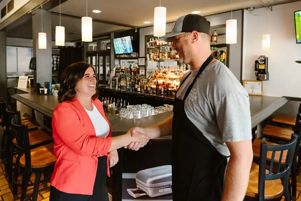A businesswoman and a bar owner shake hands in front of the bar.